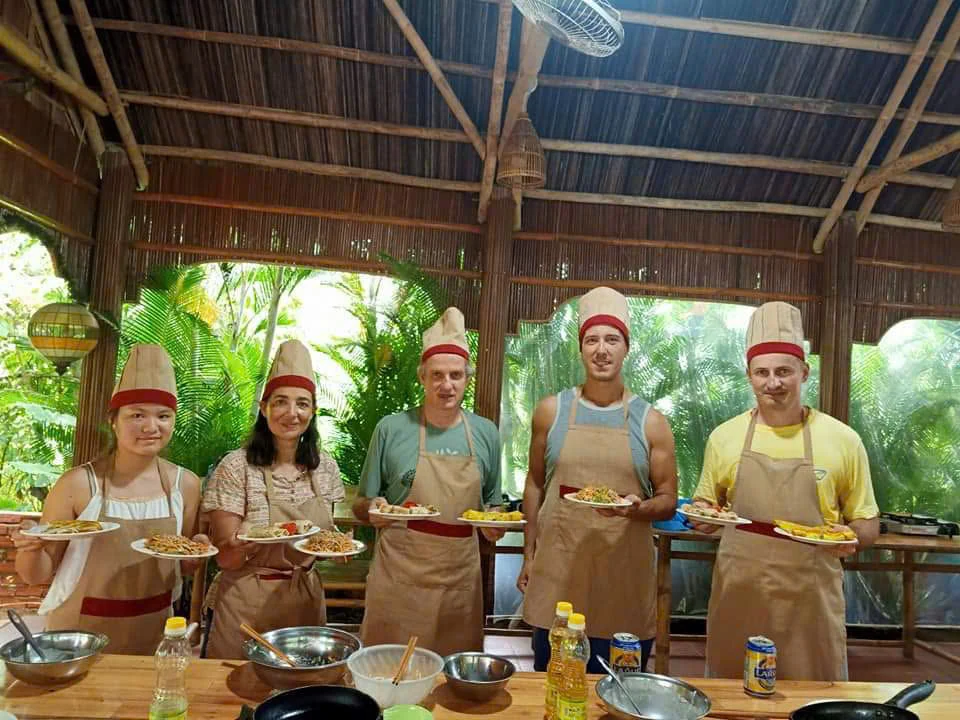 Cooking-class participants holding plates of prepared food in an open-sided kitchen.
