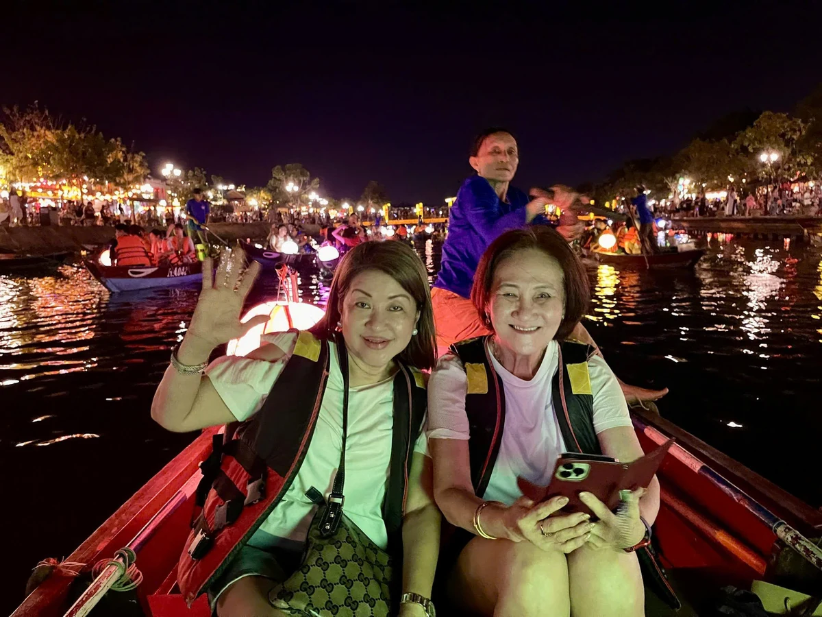 Two visitors wearing life jackets sit in a wooden boat on the lantern-lit river in Hoi An at night.