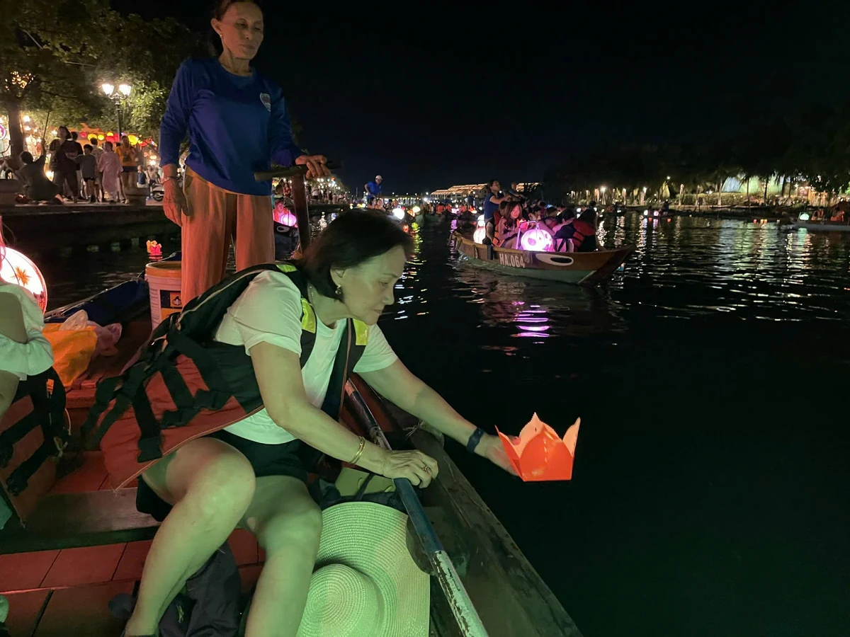 A visitor in a life jacket lowers a glowing flower-shaped paper lantern toward the river from a wooden boat in Hoi An.