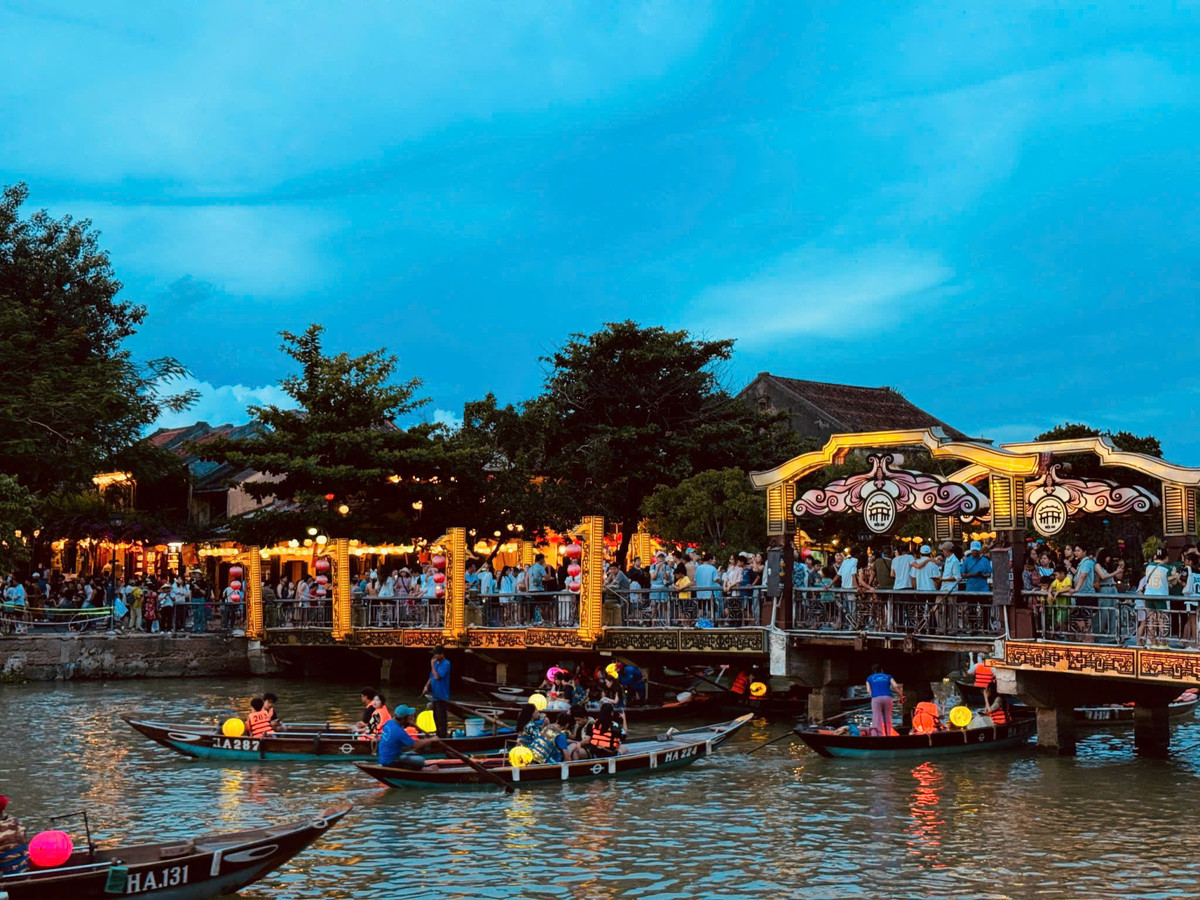 Lantern-lit wooden boats pass the illuminated An Hoi Bridge at blue hour in Hoi An.