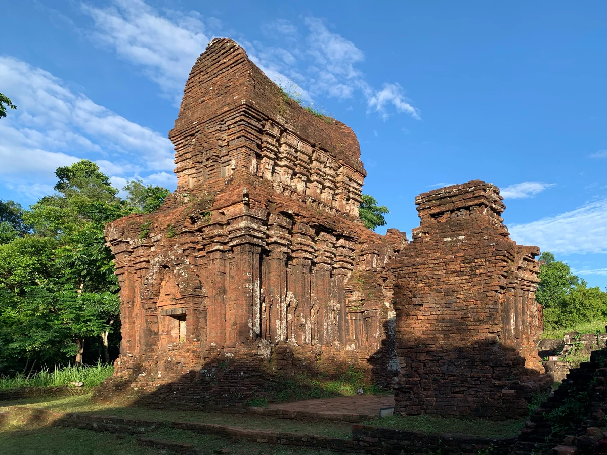 A sunlit Cham brick tower at My Son Sanctuary beneath a blue sky, with green trees around its base.