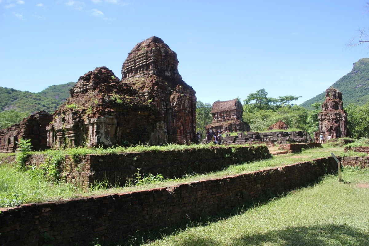 Brick Cham temples at My Son Sanctuary among grassy terraces, with forested hills and blue sky behind.