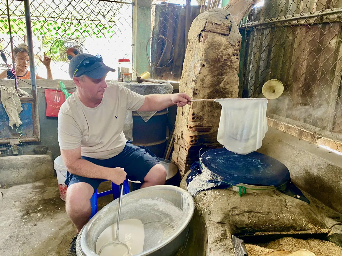 A visitor lifts a freshly steamed rice-paper sheet from the cooking cloth with a bamboo tool.