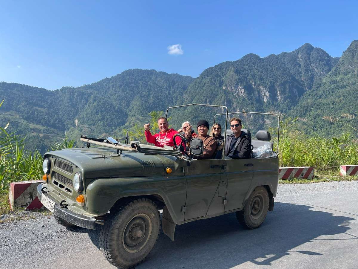 Travelers in an open-top green jeep beside a mountain road, with steep green peaks and blue sky behind them.