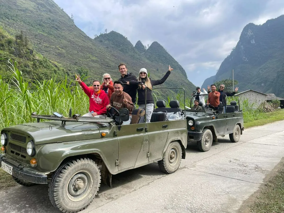 Travelers wave from two open-top green jeeps on a narrow road beneath steep rocky mountain peaks.