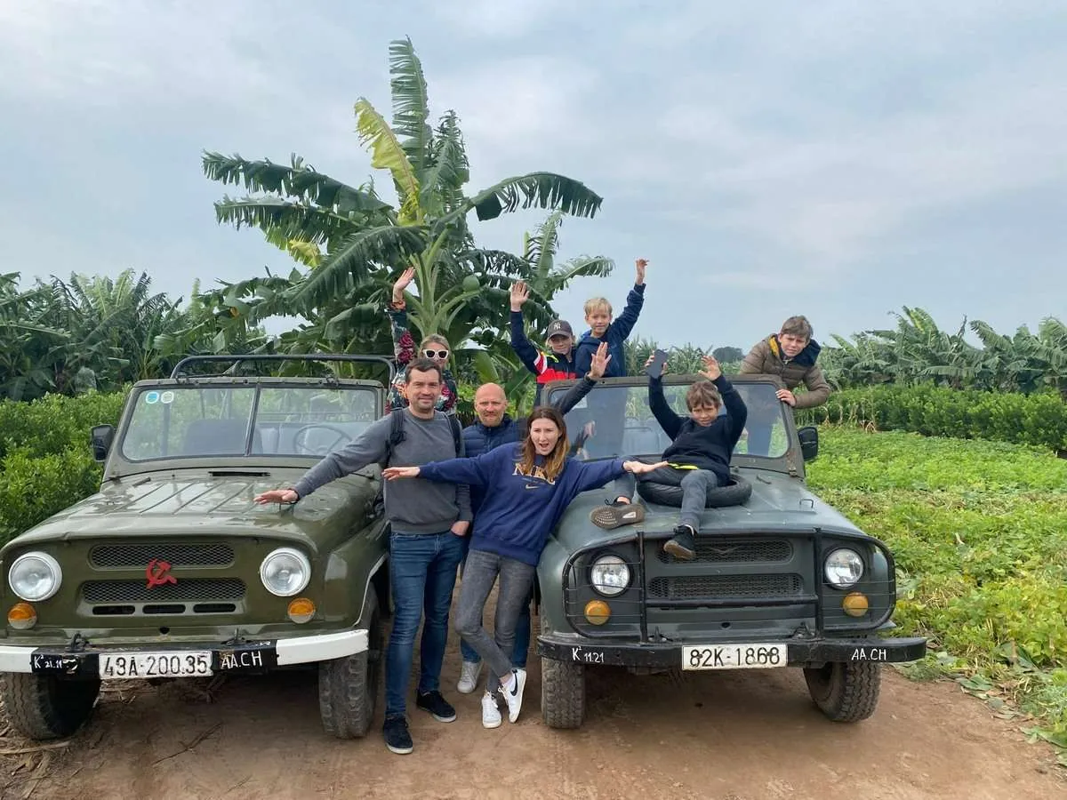 Travelers pose around two green open-top jeeps beside banana plants and green fields.