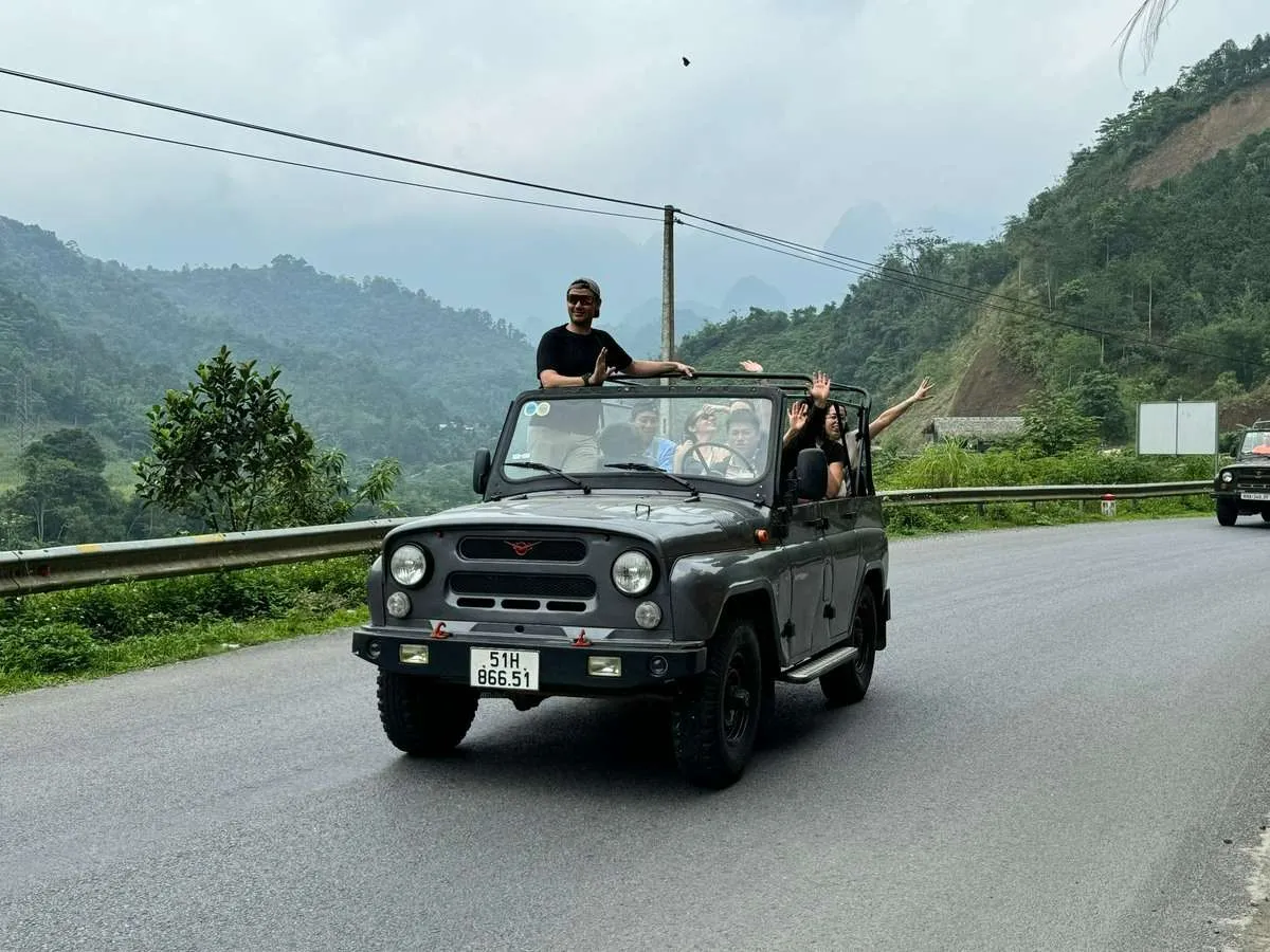 Travelers wave from an open-top jeep on a paved mountain road bordered by a guardrail and green hills.