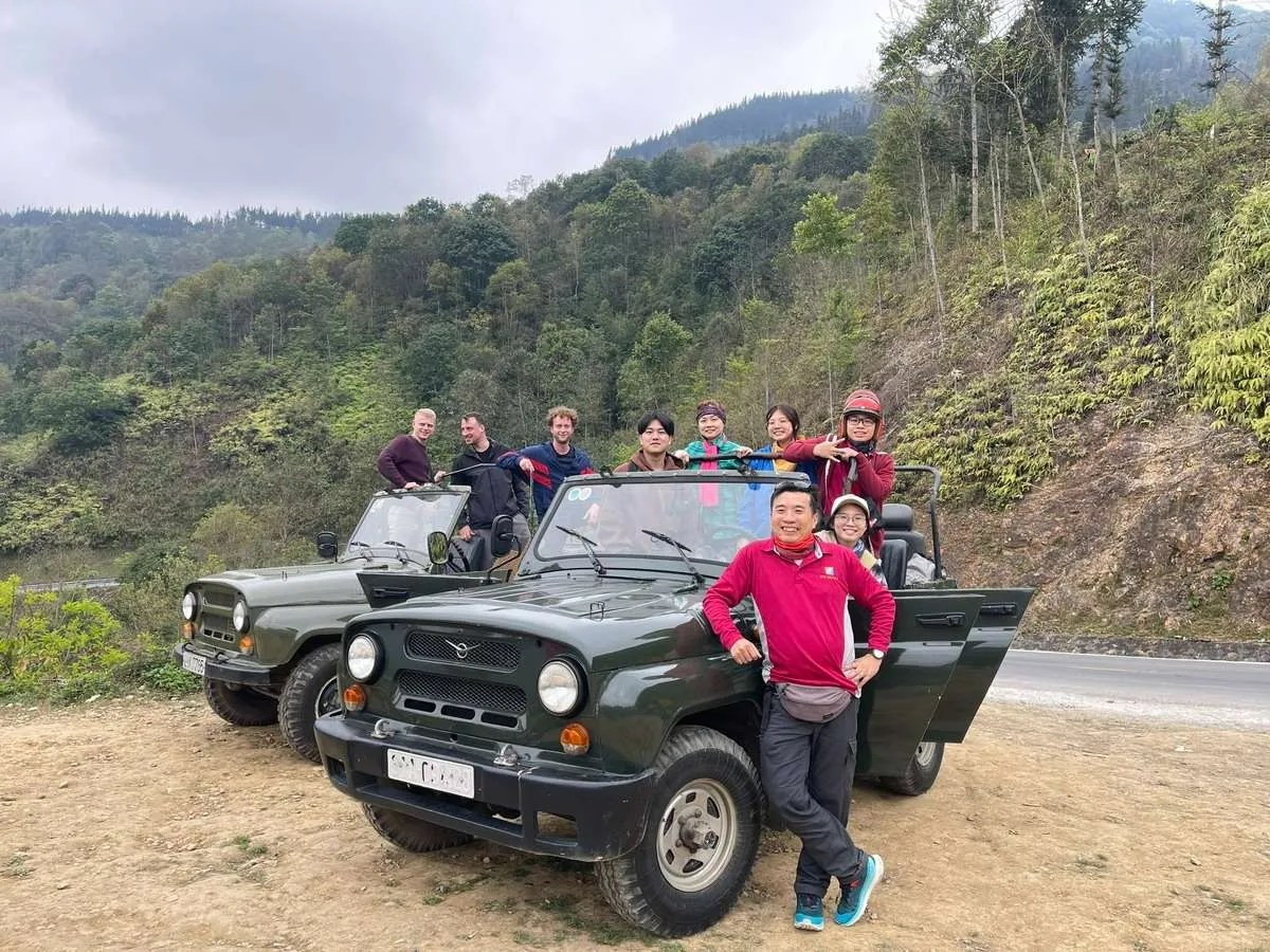 Travelers pose beside and inside two green jeeps at a roadside clearing backed by a wooded mountain slope.