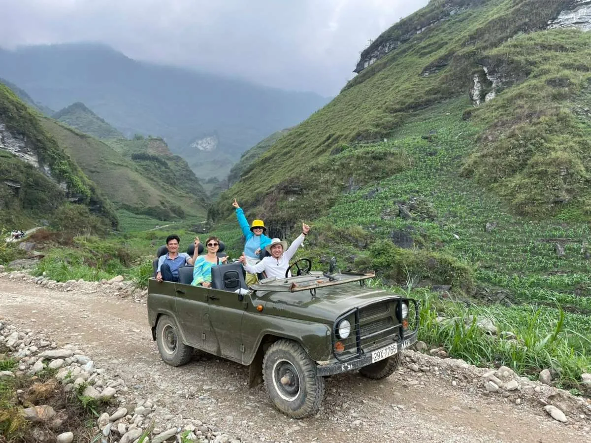 Travelers in an open-top green jeep on a rocky track between steep green mountain slopes under low cloud.