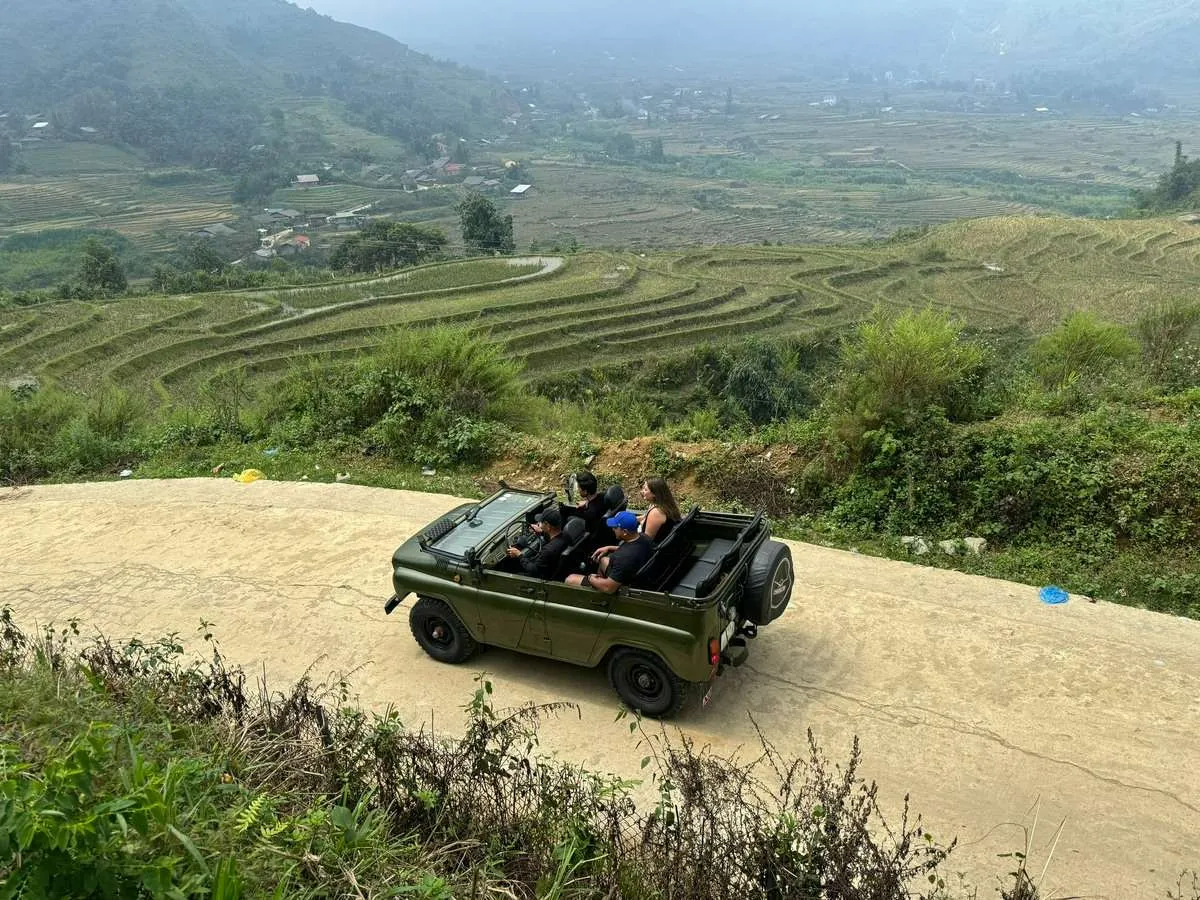 An open-top green jeep with travelers on a narrow road overlooking terraced fields and scattered valley houses.