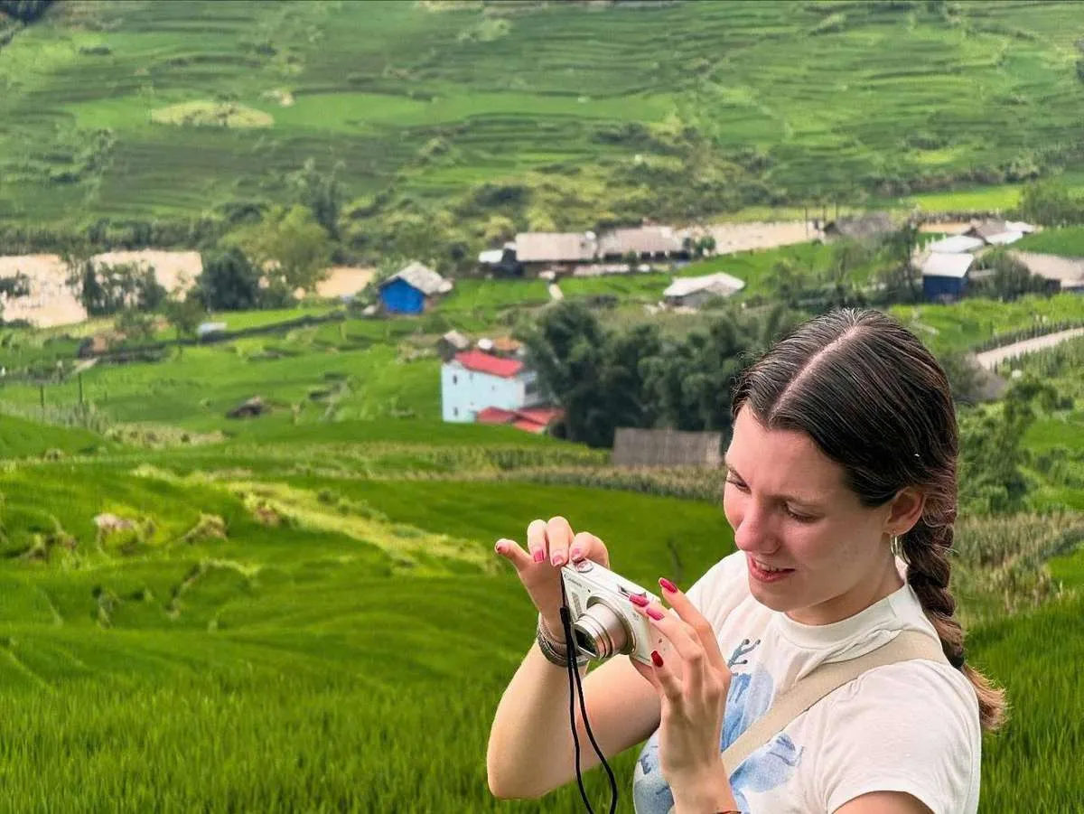A traveler looks down at a compact camera with green rice terraces and scattered houses behind them.